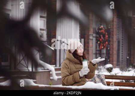 Cheerfully brunette woman drinking from flask outdoors in the city in winter. Warming up, enjoying moment, having brake over snow covered trees Stock Photo