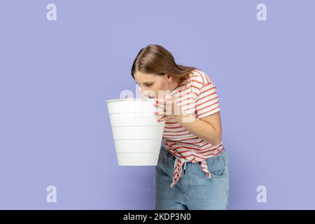 Portrait of blond woman wearing striped T-shirt feeling sudden stomach pain, suffering nausea and vomit, holding bin in hands, indigestion. Indoor studio shot isolated on purple background. Stock Photo