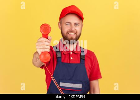 Portrait of smiling worker man holding out handset, asking call him and order service, looking friendly at camera, wearing overalls and red cap. Indoor studio shot isolated on yellow background. Stock Photo