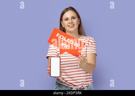 Portrait of smiling delighted blond woman wearing striped T-shirt holding red card with follow me inscription and showing white empty display of phone. Indoor studio shot isolated on purple background Stock Photo