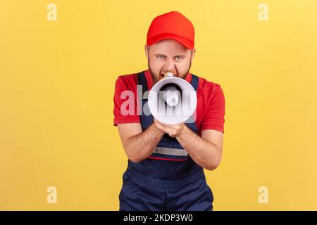 Portrait of angry worker man holding megaphone near mouth loudly speaking, screaming, making announcement, wearing blue uniform and red cap. Indoor studio shot isolated on yellow background. Stock Photo