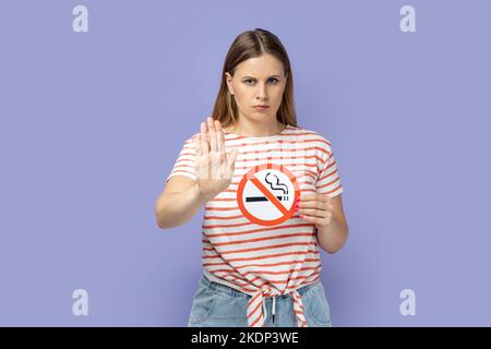 Portrait of strict attractive blond woman wearing striped T-shirt holding no smoking sign and showing stop gesture with her palm. Indoor studio shot isolated on purple background. Stock Photo