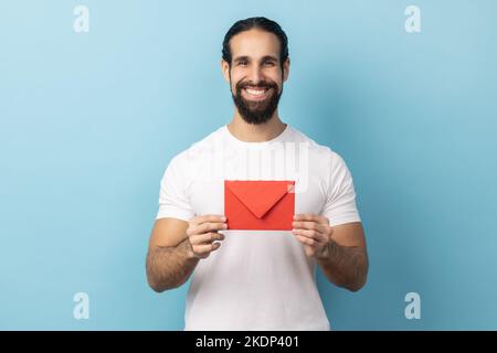 I got love letter on Valentine's day. Portrait of man with beard in white T-shirt holding letter in red envelope or greeting card and smiling joyfully. Indoor studio shot isolated on blue background. Stock Photo