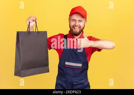 Portrait of smiling delighted bearded delivery man in blue uniform holding blag shopping bag and showing thumb up, like fast delivery service. Indoor studio shot isolated on yellow background. Stock Photo