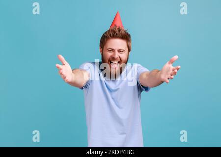 Portrait of extremely happy bearded man wearing party cone giving free hugs with outstretched hands, welcoming inviting to embrace. Indoor studio shot isolated on blue background. Stock Photo