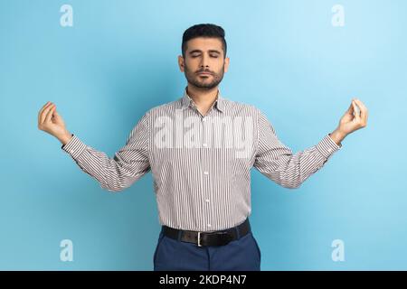 Portrait of calm relaxed young businessman with beard standing with raised arms and doing yoga meditating exercise, wearing striped shirt. Indoor studio shot isolated on blue background. Stock Photo