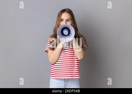 Portrait of dark haired little girl wearing striped T-shirt screaming in megaphone, speaking, announcing important information. Indoor studio shot isolated on gray background. Stock Photo