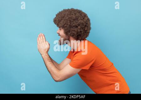 Side view of man with Afro hairstyle in orange T-shirt folding hands in prayer, closing eyes and talking to god, asking for help, expressing gratitude. Indoor studio shot isolated on blue background. Stock Photo