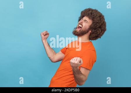 Side view of excited positive happy man with Afro hairstyle wearing orange T-shirt screaming for joy with raised fists and closed eyes, rejoicing win. Indoor studio shot isolated on blue background. Stock Photo