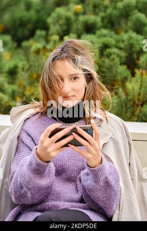 Young woman sits on bench and playing game or watching video on smartphone Stock Photo