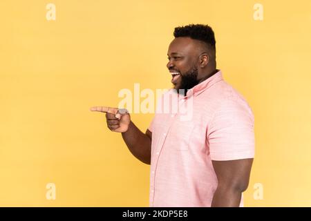 Side view portrait of extremely happy excited man wearing pink shirt standing pointing away at copy space for advertisement or promotion. Indoor studio shot isolated on yellow background. Stock Photo