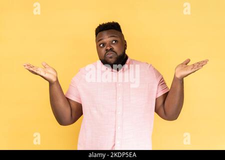 I dont know. Portrait of confused man wearing pink shirt shrugging shoulders as doesnt know answer, cant make decision, being uncertain, not sure. Indoor studio shot isolated on yellow background. Stock Photo
