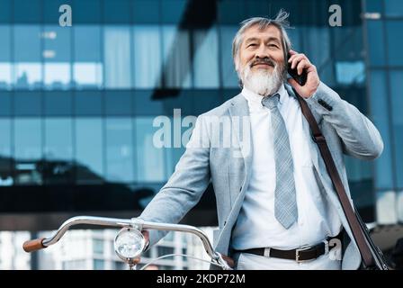 Bicycle, phone and Asian businessman in city talking, speaking and listening on phone call. Travel, eco friendly transport and mature Asian man with Stock Photo