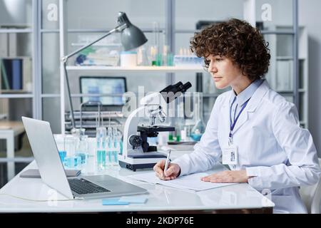 Young female scientist in labcoat making notes while sitting by workplace in front of laptop in laboratory and watching online video Stock Photo