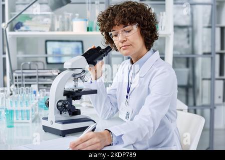Young serious woman in labcoat making notes in copybook while sitting by workplace in front of microscope and making research Stock Photo