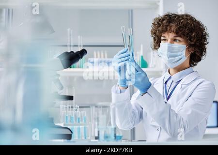 Young female virologist in protective workwear looking at two flasks with liquid substances while comparing them in laboratory Stock Photo