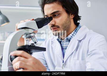 Modern male virologist in labcoat concentrating on study of new bacteria or virus while looking in microscope in clinical laboratory Stock Photo