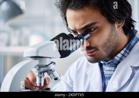 Young serious male clinician looking in microscope during research of new virus or bacteria in laboratory before creating vaccine Stock Photo