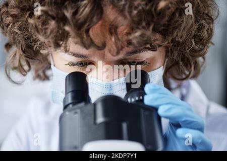Close-up of young female microbiologist in protective mask looking in microscope while studying characteristics of new virus during trial Stock Photo