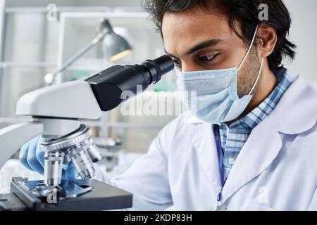 Close-up of young clinician in protective mask studying new virus or bacteria while looking in microscope by workplace in laboratory Stock Photo