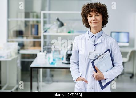 Young confident clinician with document in clipboard standing in front of camera in scientific laboratory against her workplace Stock Photo