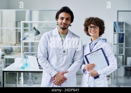 Two young clinicians or chemists in labcoats standing in front of camera in scientific laboratory against workplace and looking at you Stock Photo