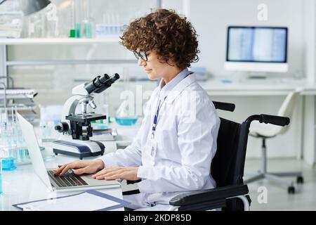 Young clinician sitting in wheelchair by desk in front of laptop and searching for online information for new scientific experiment Stock Photo