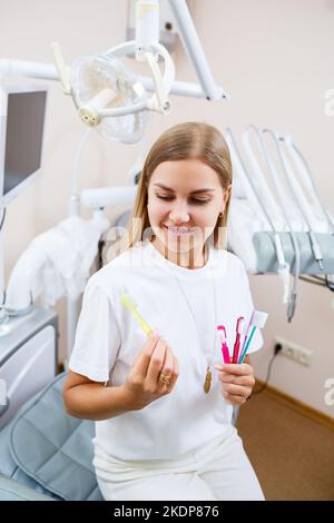 A young woman in a white t-shirt sits in a dental office, smiles and holds a toothbrush in her hands. Girl with braces shows oral care. Dentistry, den Stock Photo