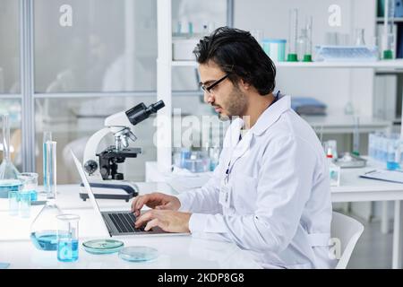 Side view of young serious laboratory worker in labcoat sitting by workplace in front of laptop and searching for online information Stock Photo