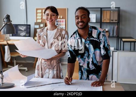 Waist up portrait of two smiling young architects working on floor plans for interior design project and looking at camera Stock Photo