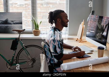 Side view portrait of young male designer using pen tablet at home office workplace and smiling Stock Photo