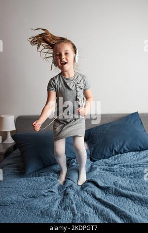 Little girl singing holding headphones cord imitating herself a real singer. Child having fun jumping dancing listening to music on bed in bedroom at Stock Photo