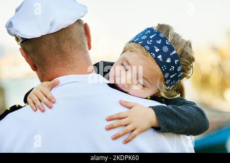 Safe and snug in daddys arms. Rearview shot of a father in a navy uniform hugging his happy little girl. Stock Photo