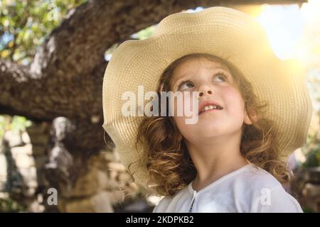 Close-up of a cute little girl wearing a rural straw hat looking up with glowing sunlight in the background Stock Photo