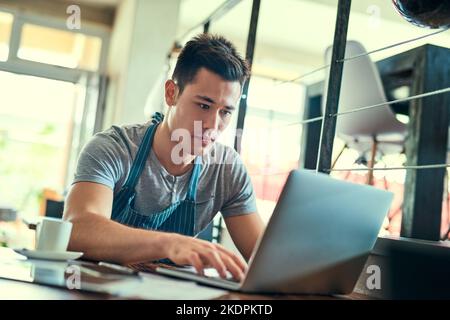 Hes got orders streaming in. a young man working on a laptop in his coffee shop. Stock Photo