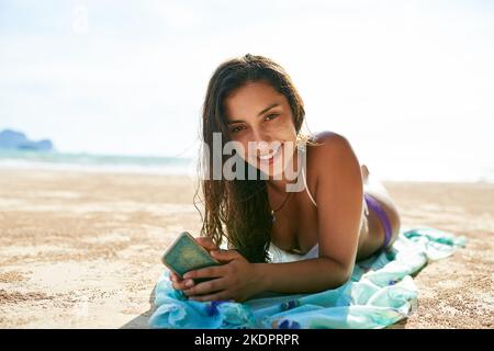 Just take the day off and go to the beach. a beautiful young woman relaxing on the beach while using her phone. Stock Photo