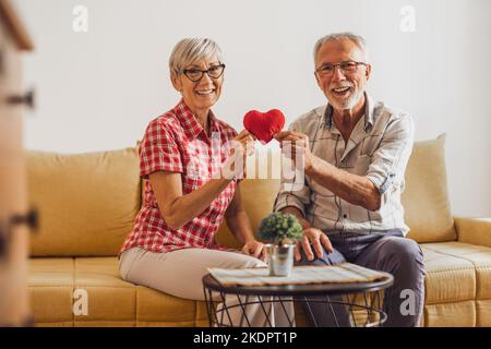 Senior couple sitting in living room and holding red heart shaped pillow. They are celebrating their anniversary. Stock Photo