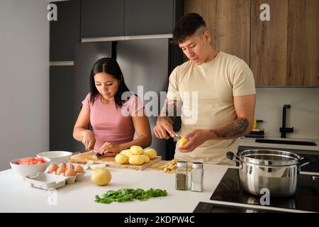 Two young latin people peeling potatoes and cutting onion at kitchen. Stock Photo