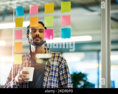 Having a caffeine boost to spark his creativity. a young man having a brainstorming session with sticky notes at work. Stock Photo
