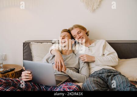 Young gay couple laughing and using laptop while resting in bed at home Stock Photo