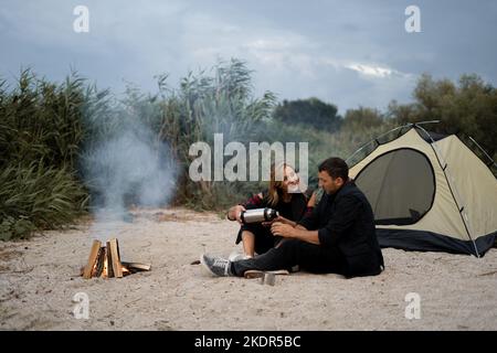 Couple of campers drinking coffee from a metal cups and a thermos while camping out in the nature in the weekend. Camping, recreation, hiking Stock Photo