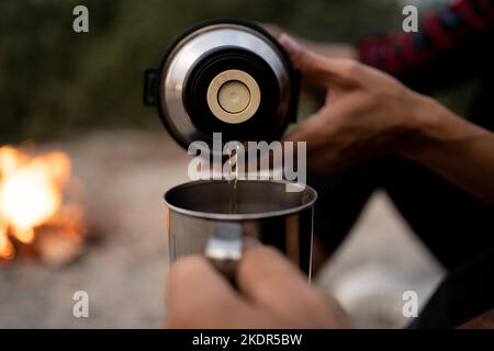 Couple of travelers drink coffee. Man pouring tea from thermos. Picnic at sea beach. Woman holding metal cup with beverage. Lifestyle moment at nature Stock Photo