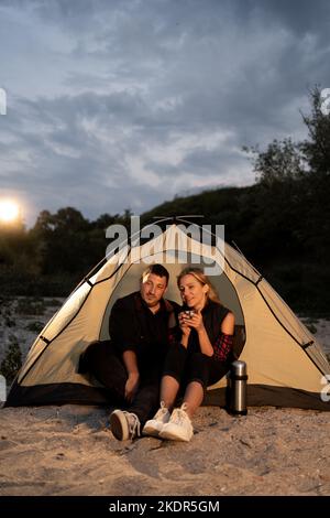 Couple of travelers set up camp on evening lake shore and relaxing in illuminated tourist tent after dinner, drinking tea. evening cloudy sky on the Stock Photo