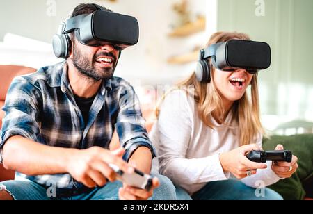 Millennial friends playing with vr glasses at home couch - Virtual reality and tech concept with engaged couple having fun on headset goggles - Genera Stock Photo