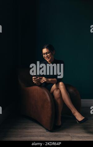 Smiling asian woman using a mobile phone while sitting on a couch in her office. Happy young woman reading a text message on her smartphone. Stock Photo