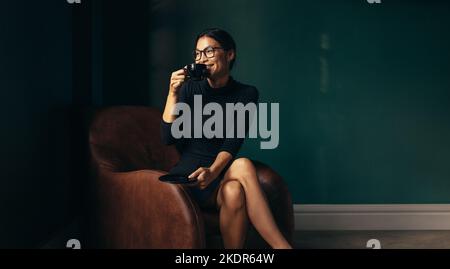 Happy asian woman sitting on sofa and drinking coffee. Beautiful businesswoman drinking coffee and looking away. Stock Photo