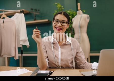 Portrait of female fashion designer sitting at desk and smiling. Woman dressmaker smiling at camera. Stock Photo