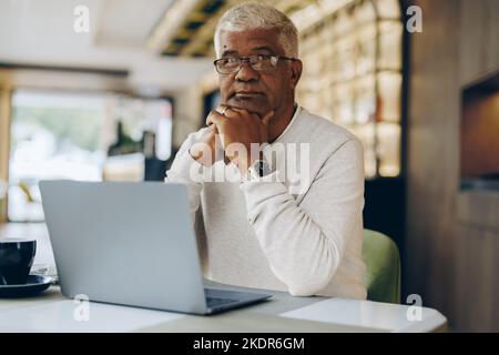 Mature businessman looking at the camera while working in a modern cafe. Senior businessman using a laptop while working remotely. Stock Photo