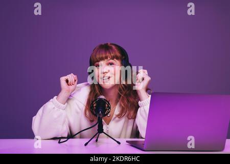 Happy young podcaster speaking into a microphone in a recording studio. Young woman hosting a live audio broadcast against a purple background. Woman Stock Photo