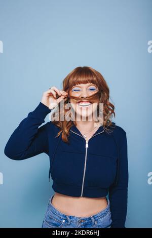 Playful young woman laughing happily while making a moustache with her hair in a studio. Cheerful young woman having fun while standing against a blue Stock Photo